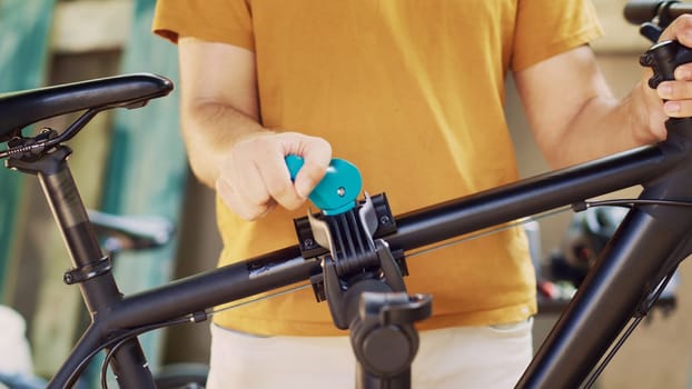 Close-up of sports-loving male grasping and securing damaged bike for adjustment on repair-stand. Caucasian person doing maintenance on bicycle while using tools in yard during summer.