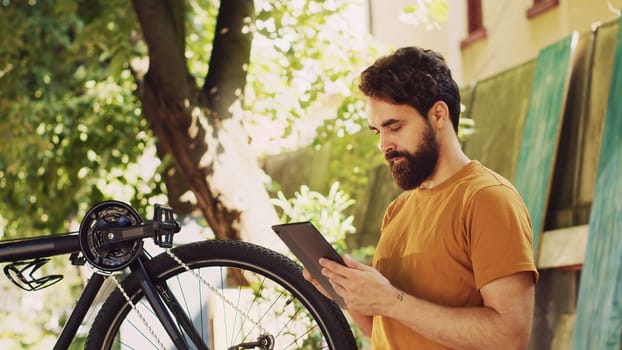 Determined active man examining and fixing his bicycle while using his digital tablet for guidance. Young sporty dedicated male cyclist browsing the internet to maintain bicycle in yard.