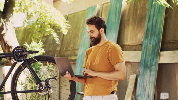 Male caucasian cyclist utilizing laptop to reasearch for online repair guides while repairing broken bicycle pedal. Healthy young man upkeeping bike by surfing the internet on minicomputer.