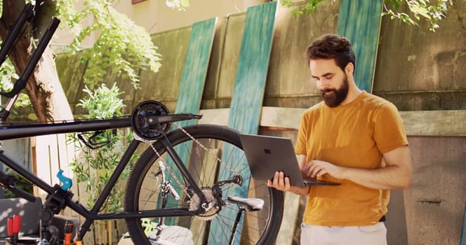 Sports-loving healthy man using laptop for enhancing safety of bike components for leisure cycling. Active male cyclist holding minicomputer for instructions to help repair damaged bicycle wheel.