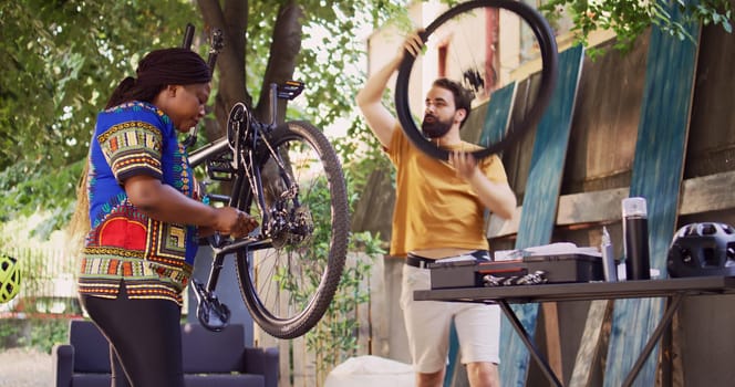 Young multiracial pair fixes broken bike with professional equipments. Caucasian man dismantling wheel while black woman adjusts chains and pedals for yearly bicycle repair and maintenance.