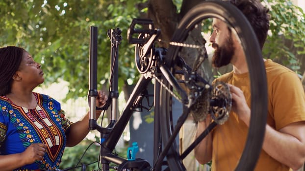 Fit and dedicated multiracial pair inspecting damaged bicycle for maintenance using professional equipment. Boyfriend mending bike pedal and talking with girlfriend who is removing broken wheel.