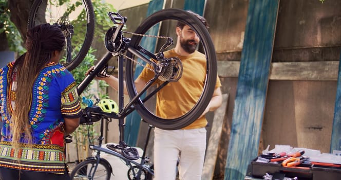 Young sports-loving multiracial couple repairing broken bicycle using professional equipment. Caucasian male loosening and dismantling damaged bike tire while black woman prepares work tools.