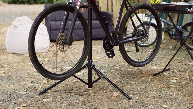 In yard, modern black bike seen stationed on repair-stand for fine-tuning and adjusting of components. Damaged bicycle parts outside waiting for repair with specialized work tools.