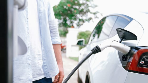 Modern eco-friendly man recharging electric vehicle from home EV charging station. EV car technology utilized for home resident to future environmental sustainability. Synchronos