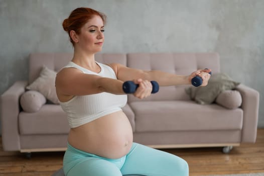Pregnant woman doing exercises with dumbbells while sitting on a fitness ball at home