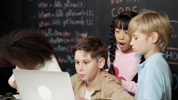 Closeup of boy using laptop programing engineering code and writing program while group of smart diverse student standing surrounded by friend in STEM technology classroom at blackboard. Erudition.