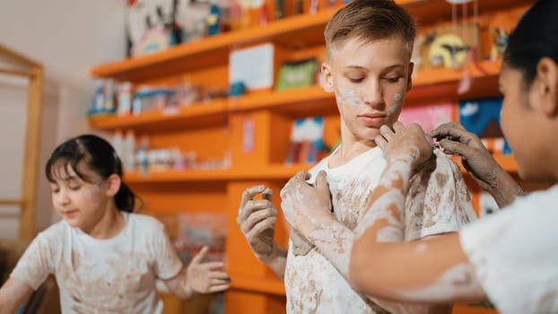 Smart caucasian teenager and happy friend with mixed races put clay on body in art lesson. Diverse highschool student playing and modeling cup of clay at workshop in creative activity. Edification.