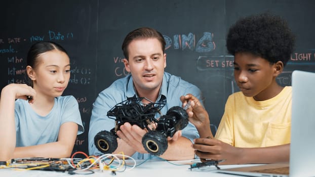 Smart teacher inspect car model construction to diverse student. Highschool children with mixed races listening mentor explain robotic model system at table with laptop and wires placed. Edification.