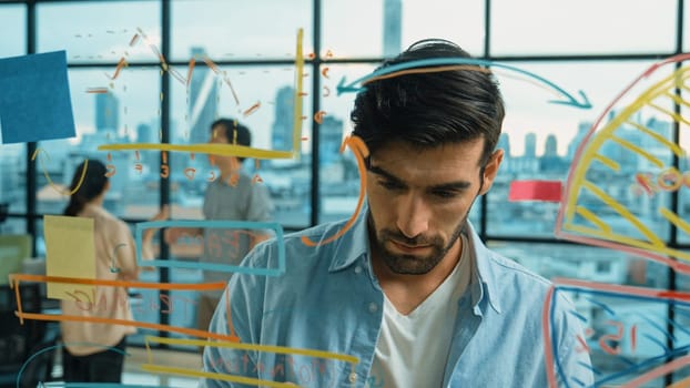 Professional businessman uses sticky notes at glass wall with graph to brainstorming idea while multicultural businesspeople discussing about marketing ideas at office with city view. Tracery