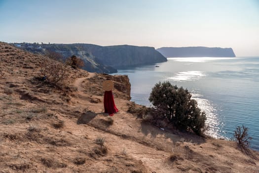 A woman in a red dress stands on a rocky cliff overlooking the ocean. She is wearing a straw hat and she is enjoying the view