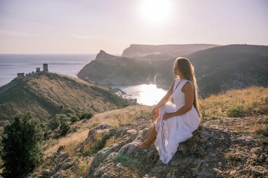 A woman in a white dress sits on a rock overlooking a body of water. The scene is serene and peaceful, with the woman enjoying the view and the calmness of the surroundings