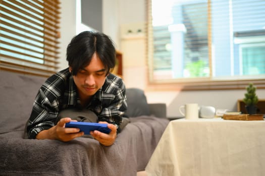 Relaxed young Asian man lying on sofa and checking social media on smartphone.