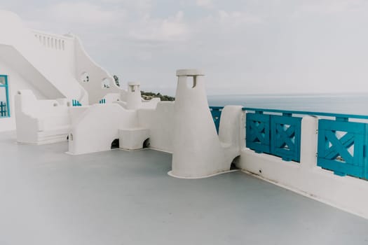 A white building with blue trim and a blue balcony. The balcony overlooks the ocean. The building is empty and the sky is cloudy