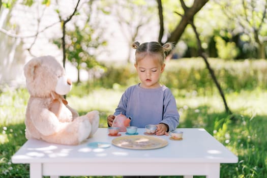 Little girl stands at a table in the garden and plays tea party with a teddy bear. High quality photo