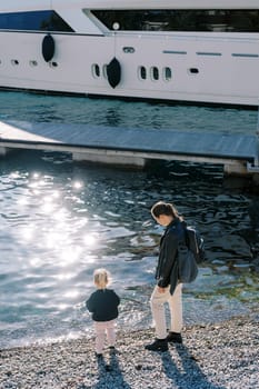 Mother and a little girl stand on a pebble beach near a moored yacht and look at the water. Back view. High quality photo