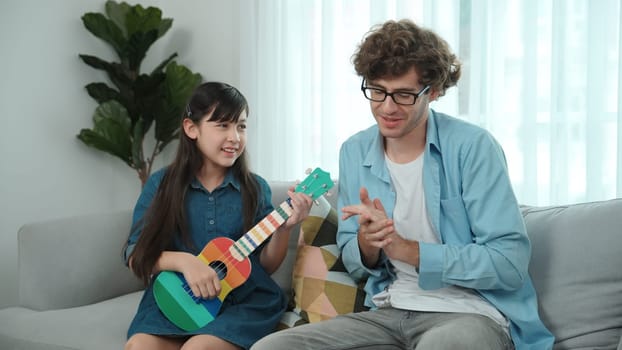 Back view of happy father looking at smart daughter playing ukulele warmly and singing song. Happy dad motivated and encouraged american school girl enjoy playing guitar. Family recreation. Pedagogy.