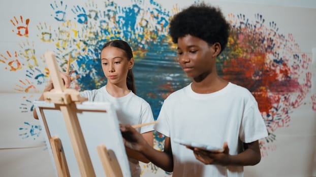 Diverse happy children paint canvas together with colorful stained wall. Smart multicultural student drawing together in art lesson while wearing white shirt. Creative activity concept. Edification.