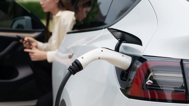 Young woman use smartphone to pay for electricity at public EV car charging station green city park. Modern environmental and sustainable urban lifestyle with EV vehicle. Expedient