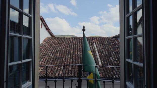 Brazilian flag on a balcony with open doors, building's roof in the background.