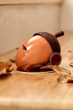 Tempting chocolate acorn shaped cake in glossy hazelnut praline ganache presented on wooden surface surrounded by dried oak leaves. Artisan dessert inspired by bounties of nature
