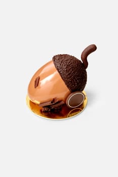 Delicate acorn shaped cake covered in glossy hazelnut praline ganache with peanut cremeux filling and dark chocolate cap and stem, served on golden cardboard on white background. Signature dessert