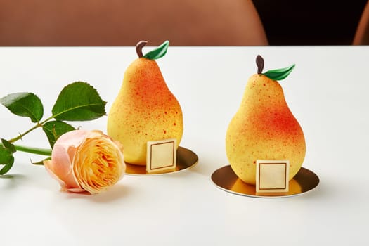 Two exquisite pear shaped pastries with creamy mousse and fruit confit on golden cardboards accompanied by delicate rose, evoking sense of gourmet luxury. Collections of signature handmade desserts