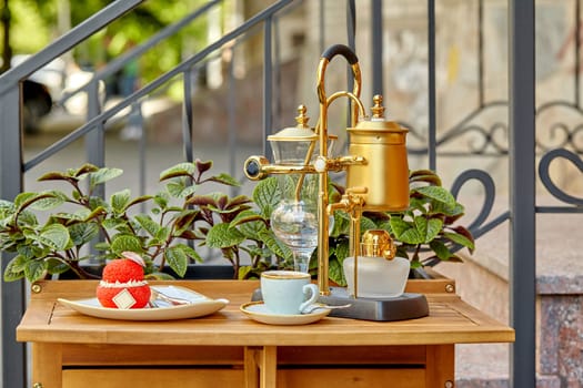 Cozy setting on outdoor cafe featuring wooden table served with vintage luxurious golden coffee siphon brewer and artisan red choux au craquelin pastry filled with custard