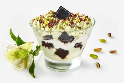 Exquisite pistachio-flavored layered dessert garnished with chocolate and pistachio nuts, accompanied by delicate greenish flower on white background