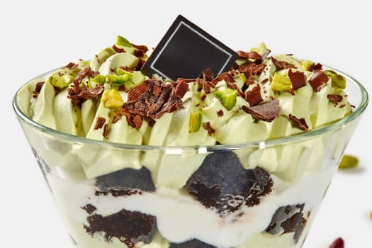 Closeup of delicious layered trifle with pieces of brownie, pistachio ice cream and whipped cream garnished with chocolate shavings and nuts served in dessert glass on white background