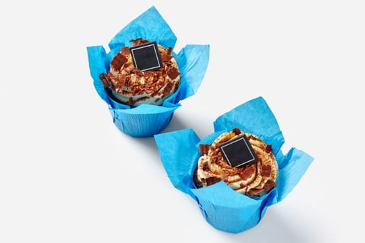 Two freshly baked cupcakes topped with whipped cream swirls sprinkled with cocoa and chocolate shavings in bright blue paper wrappers on white background. Popular confectionery products