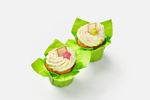 Two vanilla cupcakes topped with whipped cream decorated with colorful chocolate flowers in bright green paper wrappers on white background. Popular confectionery products