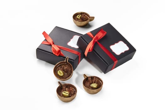 Gourmet chocolate candies crafted in shape of miniature cups of espresso with creamy filling and golden coffee beans, presented in stylish black gift boxes with vibrant red ribbons on white background