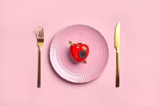 Red heart-shaped strawberry mousse dessert with glossy icing, chocolate and golden arrow elegantly placed on textured porcelain plate with gold cutlery on pink background. Romantic declaration of love