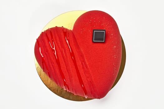 Top view of velvety vibrant red heart-shaped mousse cake decorated with glossy icing and chocolate on gold cardboard, isolated on white background. Romantic delicious gift for Valentines Day