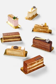 Slices of variety of exquisite cakes with velvety chocolate sponge, light creamy and berry mousses arranged on golden serving cardboards against white background. Popular baked sweets