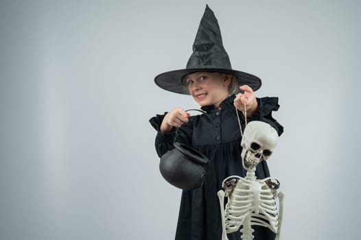 Portrait of a little Caucasian girl in a witch costume holding a cauldron and a skeleton on a white background