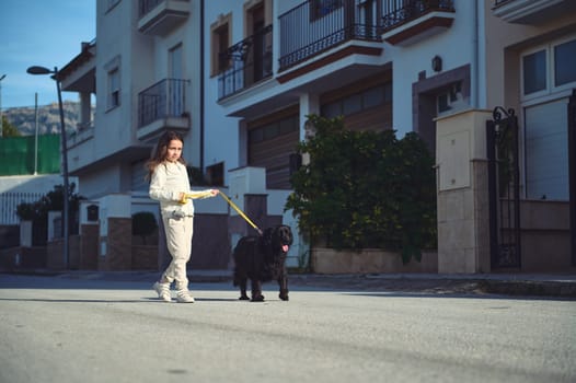 I'm taking my pet for walk. Cute child girl walking her dog on leash. Full length portrait of a happy kid in white sportswear, enjoying a morning walk with her pedigree black purebred cocker spaniel