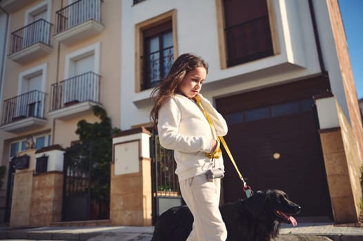 European adorable little girl taking her dog for a walk on leash on the city street on a sunny day, dressed in white sportswear. Lovely kid walking her cocker spaniel outdoors