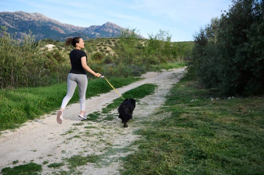 Full length rear portrait of a multi ethnic young woman running while walking her dog on leash on the nature outdoors. People. Nature. Domestic animals. Playing pets