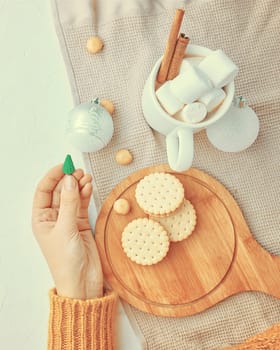 Woman in a yellow sweater drinks hot cocoa with marshmallow and cinnamon rolls in a white mug and cookies and a figure of a Christmas tree