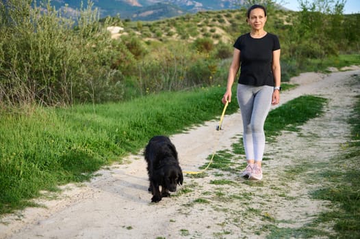 Full size shot beautiful happy smiling woman walking her dog in mountains nature outdoors. Front view of athletic lady in sportswear, walking her pedigree cocker spaniel while performing morning jog