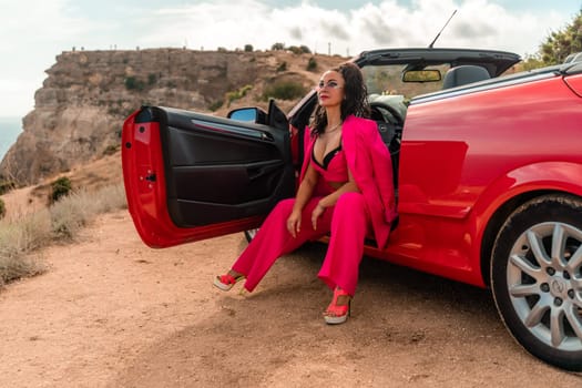 A woman in a pink suit sits in the open door of a red convertible. The scene is set on a rocky hillside overlooking the ocean. The woman is posing for a photo