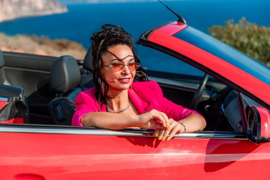 A woman in a pink jacket is sitting in a red convertible. She is wearing sunglasses and has her hand on her hip. Scene is relaxed and carefree