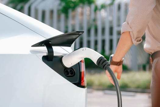 Young man put EV charger to recharge electric car's battery from charging station in city commercial parking lot. Rechargeable EV car for sustainable environmental friendly urban travel. Expedient