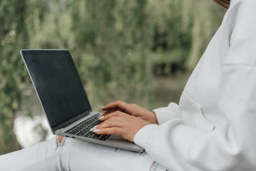 A woman is sitting on a bench and typing on a laptop. She is wearing a white sweater and has her hands on the keyboard. Concept of productivity and focus