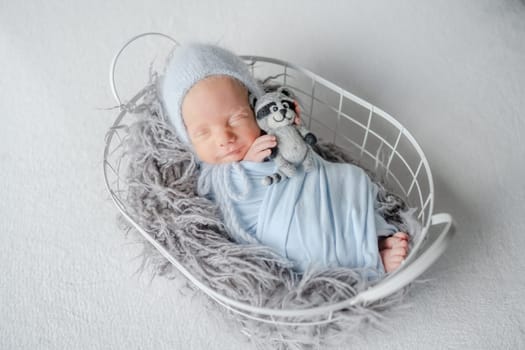 Newborn Baby In Grey Wrap Sleeps In Metal Basket During Studio Photoshoot