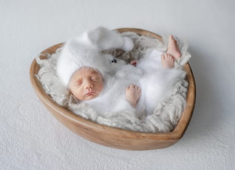 Newborn Baby In White Jumpsuit Sleeps In Wooden Heart-Shaped Bowl During Studio Photoshoot
