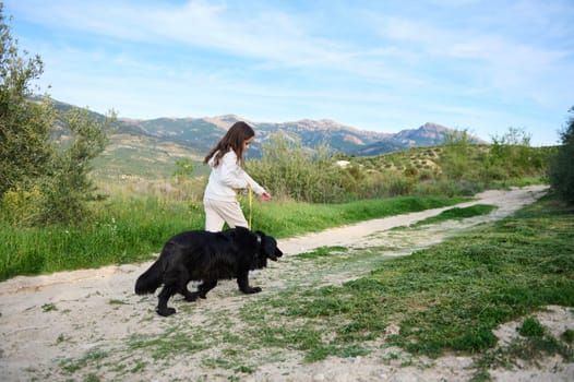 Adorable European child girl walking her pedigree dog, a black purebred cocker spaniel in the hills mountains nature outdoors on a sunny day. People. Nature and playing pets concept. Rear view