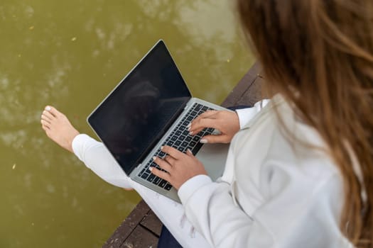 A woman is sitting on a dock with a laptop open in front of her. She is typing on the keyboard and she is focused on her work. Concept of productivity and concentration